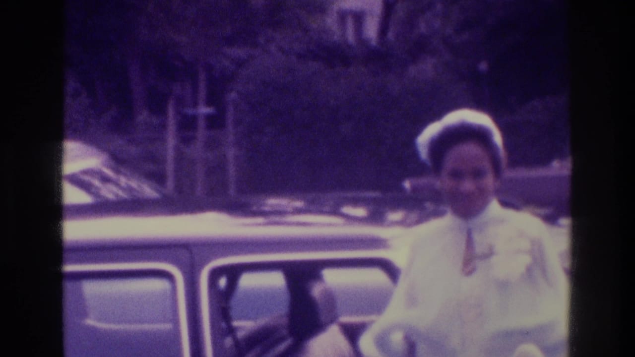 The bride, wearing a white dress and hat, stands beside a dark car in the 1975 Atlanta wedding home movie.