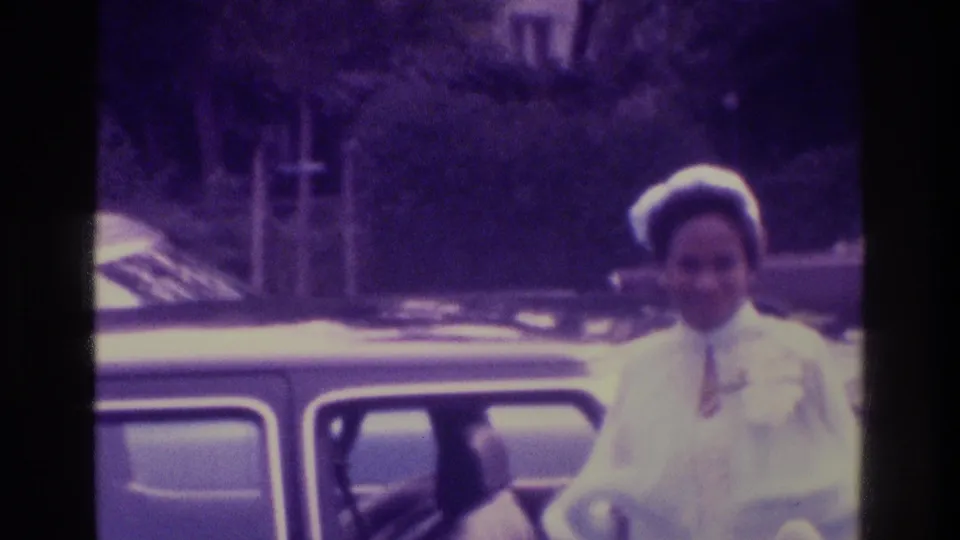 The bride, wearing a white dress and hat, stands beside a dark car in the 1975 Atlanta wedding home movie.
