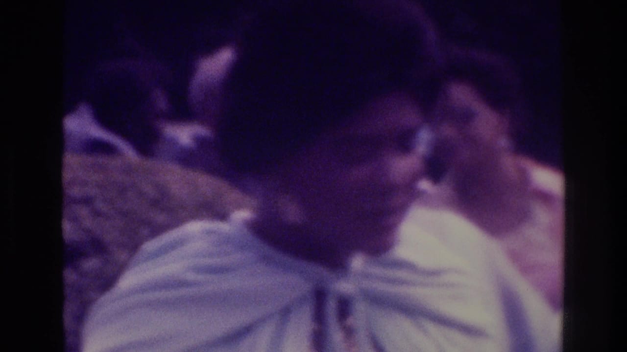 Dark-haired woman in a light dress appears close to camera among other adults.