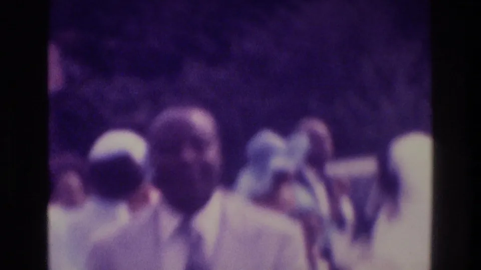 Grainy 1975 Atlanta 8mm frame showing suited adults and others in light clothing gathered outdoors, with faces softened by motion and focus.