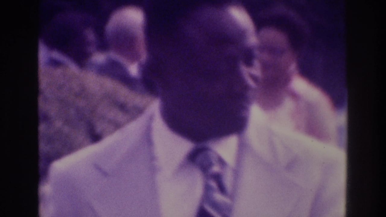 1975 Atlanta home-movie frame showing a man in a light suit and tie near the camera, with a soft, purple-tinted crowd in the background.