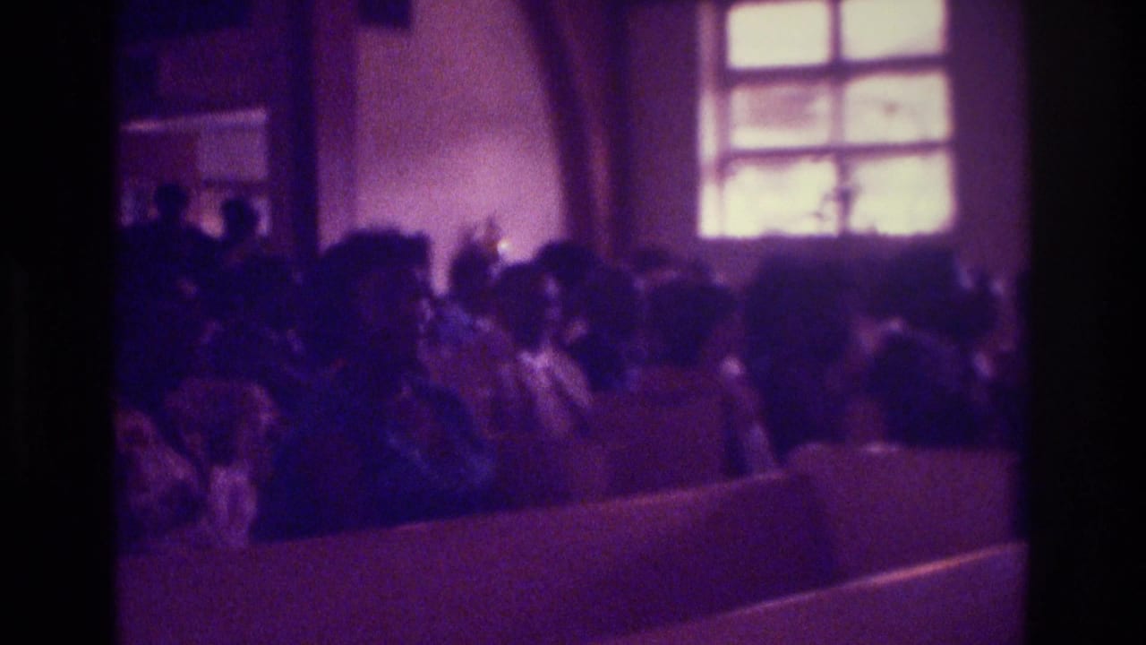 1975 Atlanta home-movie frame showing seated church attendees in pews, softened by low light and purple film cast.