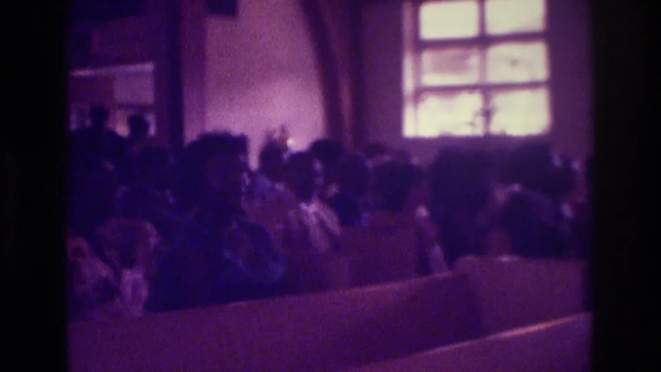 1975 Atlanta home-movie frame showing seated church attendees in pews, softened by low light and purple film cast.
