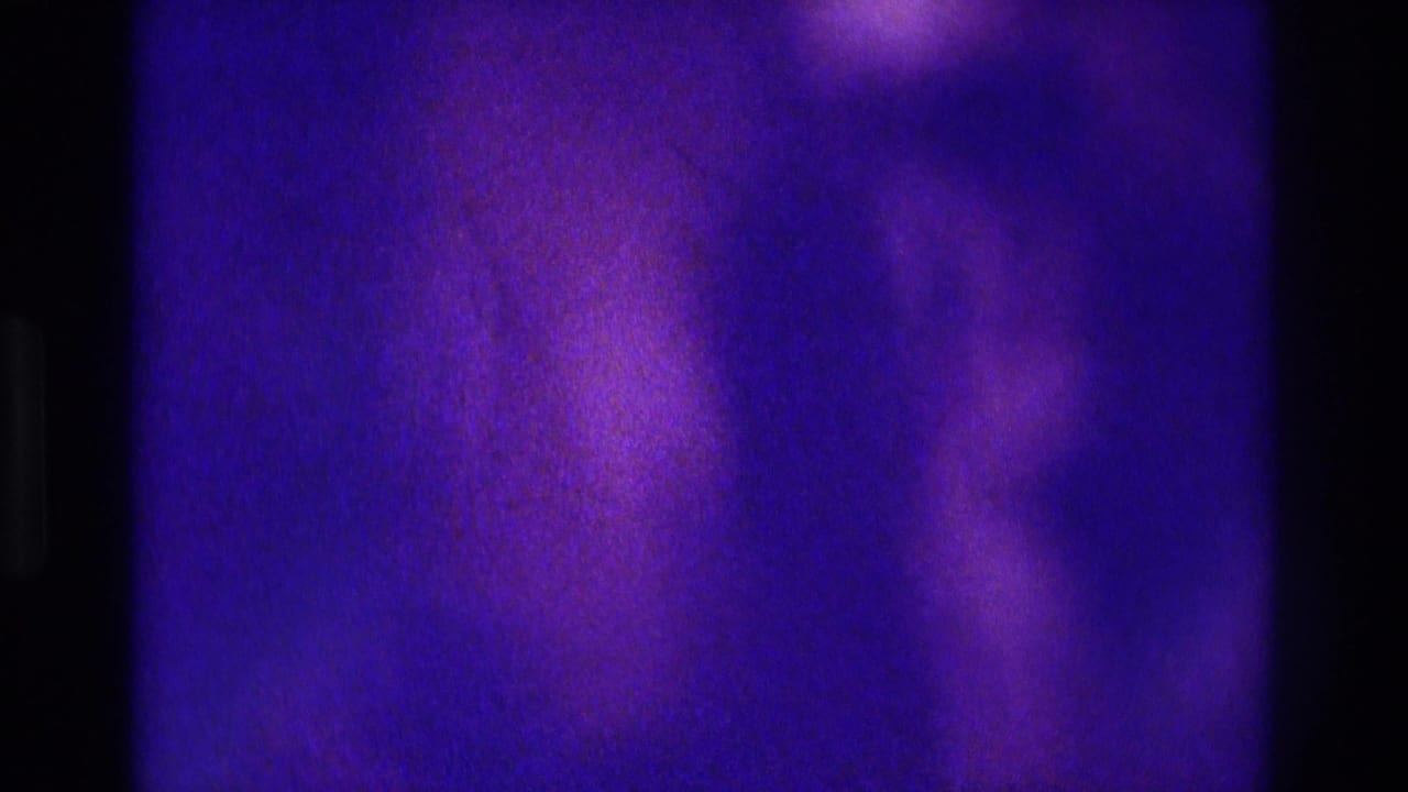 Low-light 8mm frame from the 1975 Atlanta party footage, mostly abstract purple-blue grain with no clearly identifiable people.