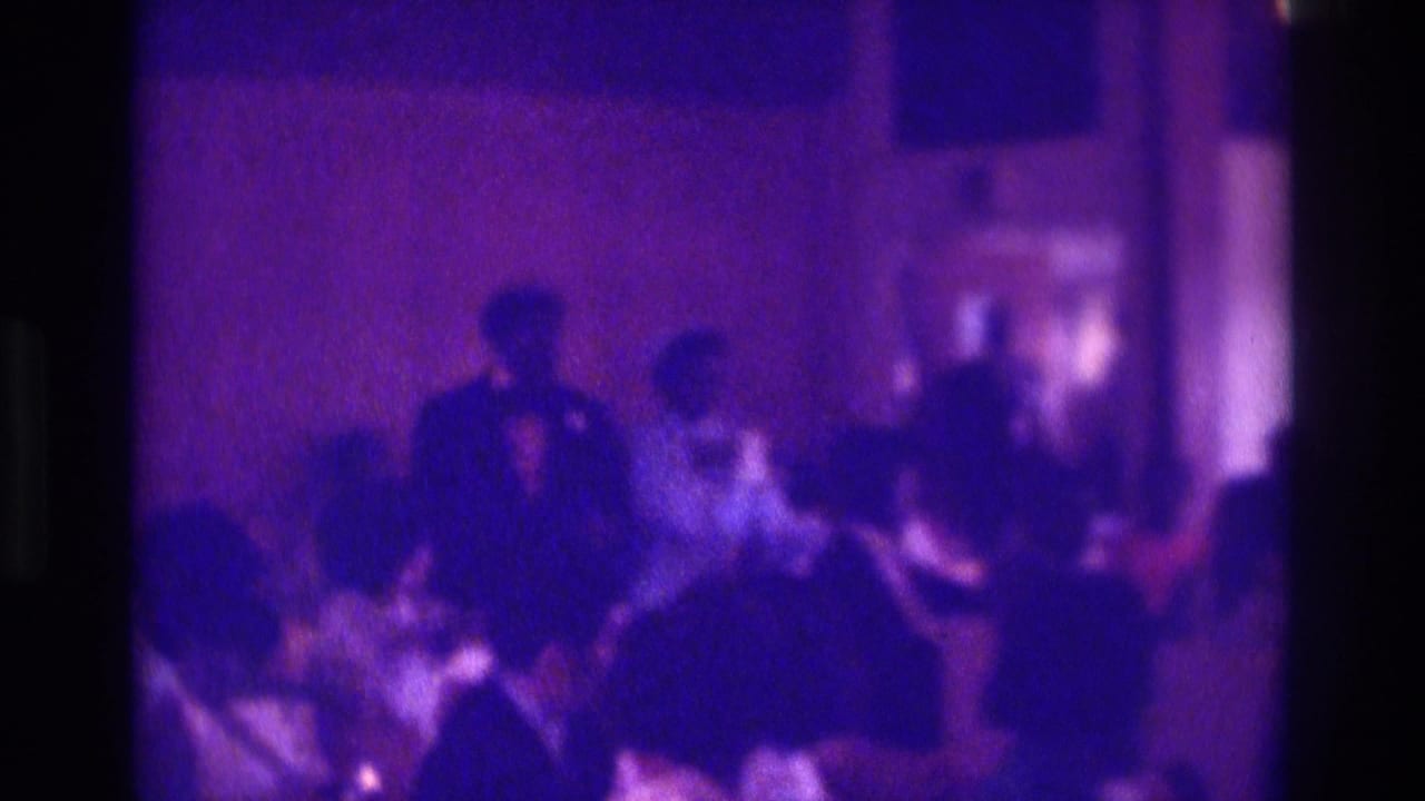1975 Atlanta home-movie frame of a Black couple moving down an indoor wedding aisle with seated guests nearby.