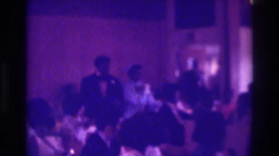 1975 Atlanta home-movie frame of a Black couple moving down an indoor wedding aisle with seated guests nearby.