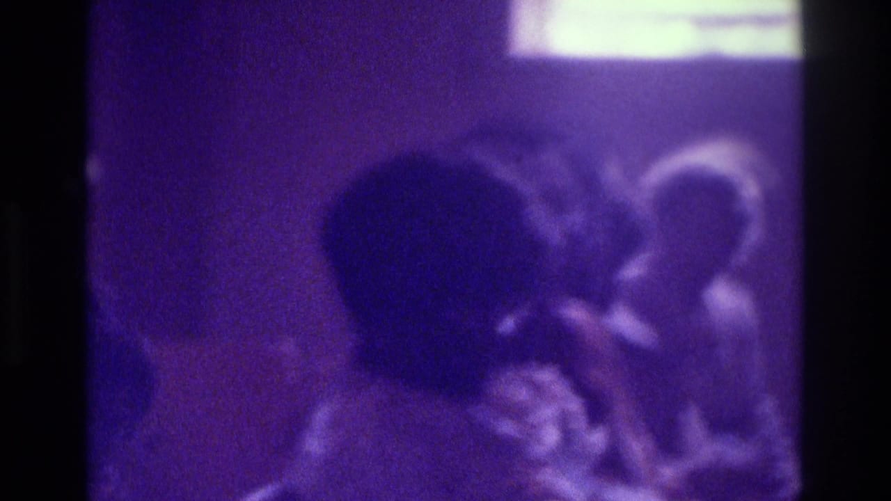 1975 Atlanta 8mm frame of indistinct seated people in a dim purple-tinted interior with a bright light area above.