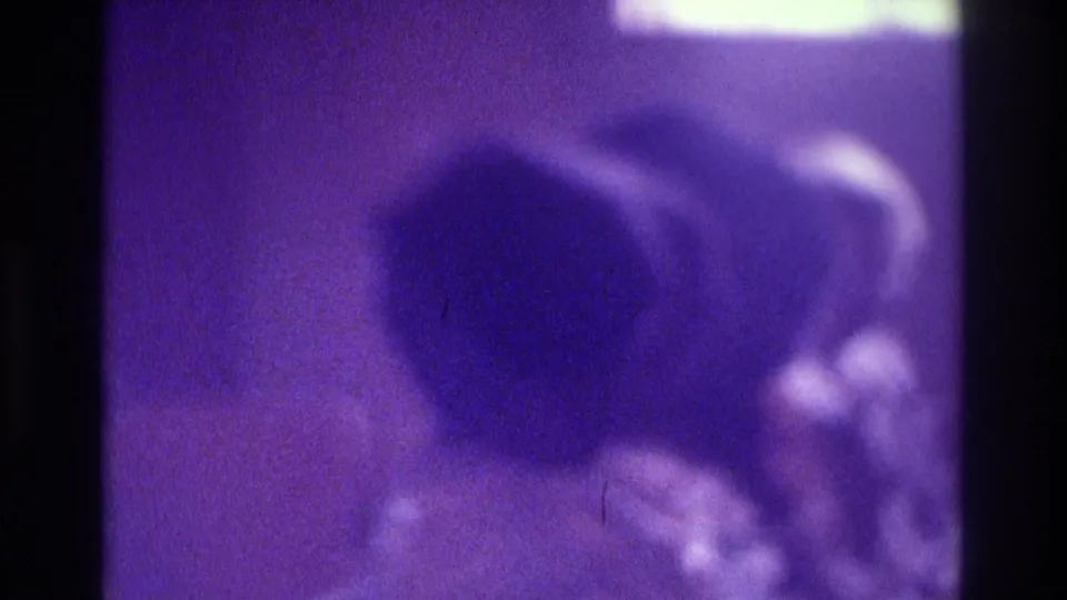 1975 Atlanta home-movie frame with a dark-haired seated figure and another partial figure in a purple, dim interior.