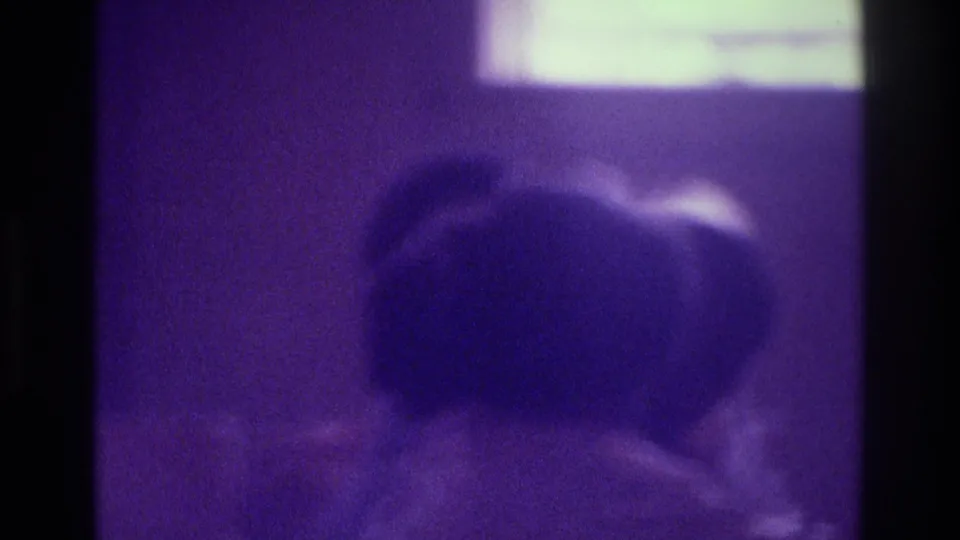 Purple-tinted 8mm frame from 1975 Atlanta showing indistinct seated figures below a bright rectangular light source.