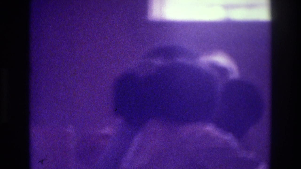 1975 Atlanta 8mm frame with blurred indoor figures, heavy grain, dark borders, and a strong purple-blue color cast.