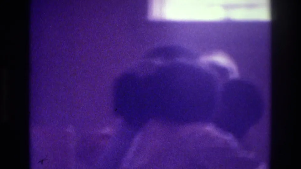 1975 Atlanta 8mm frame with blurred indoor figures, heavy grain, dark borders, and a strong purple-blue color cast.