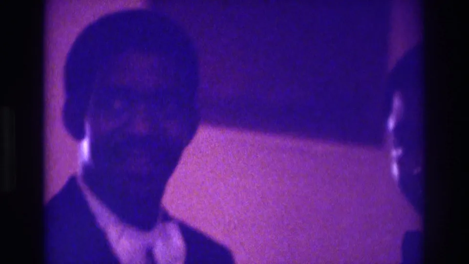 1975 Atlanta 8mm frame of an African American man in a dark suit and light shirt indoors, partly shadowed and softly focused.