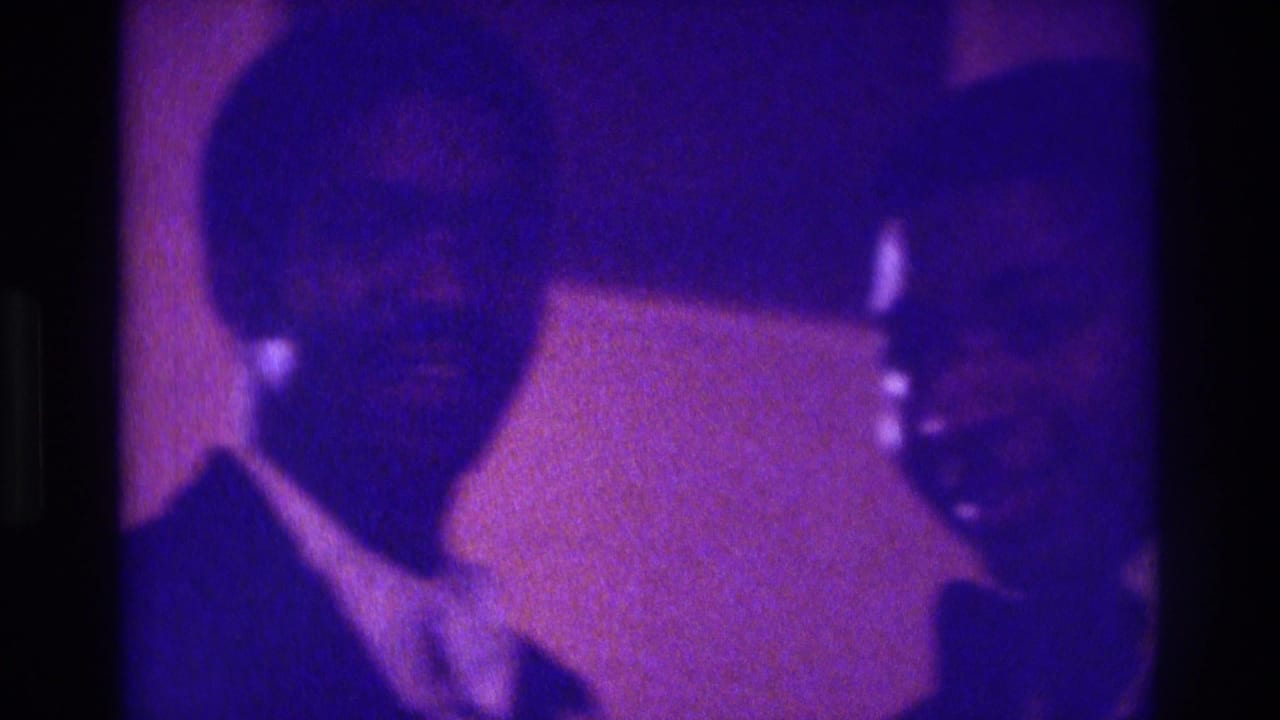 In Atlanta 1975 home-movie footage, two formally dressed men appear close to the camera in grainy purple-blue event light.