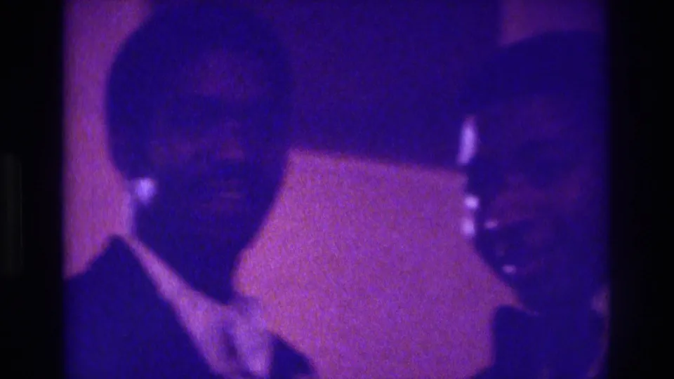 In Atlanta 1975 home-movie footage, two formally dressed men appear close to the camera in grainy purple-blue event light.