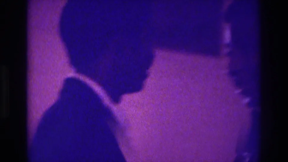 A formally dressed man appears in profile beside another person, filmed in soft-focus purple-blue light in Atlanta 1975 footage.