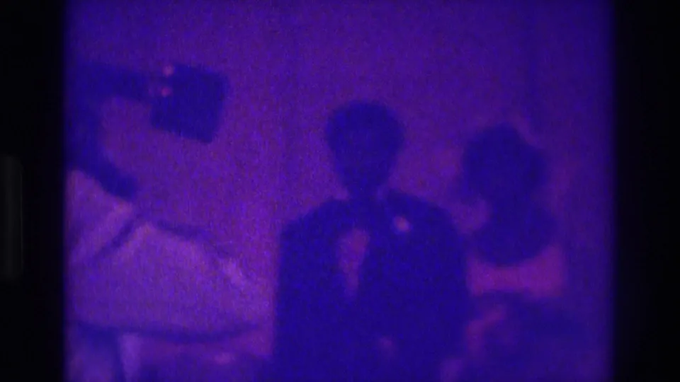 A suited man stands mid-frame among other guests, with dim formal-event lighting and heavy 8mm grain.