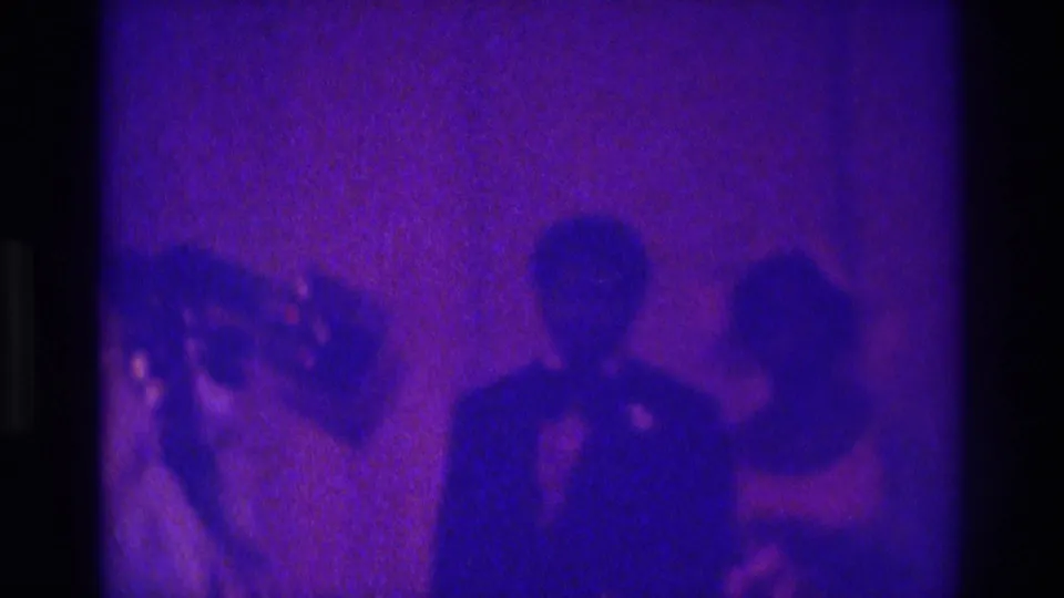A formally dressed man faces the camera with another person beside him, captured in grainy Atlanta 1975 home-movie footage.