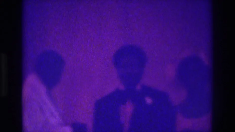 Dim 1975 home-movie frame with a suited figure and two nearby guests, softened by grain and purple color distortion.
