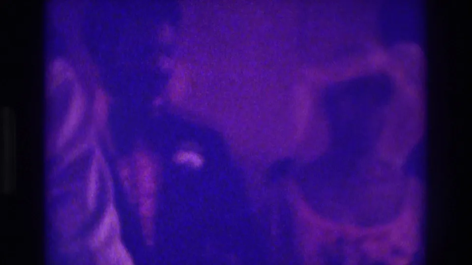 Atlanta 1975 8mm frame showing a veiled figure, suited person, and guest in a dim event room with heavy grain.