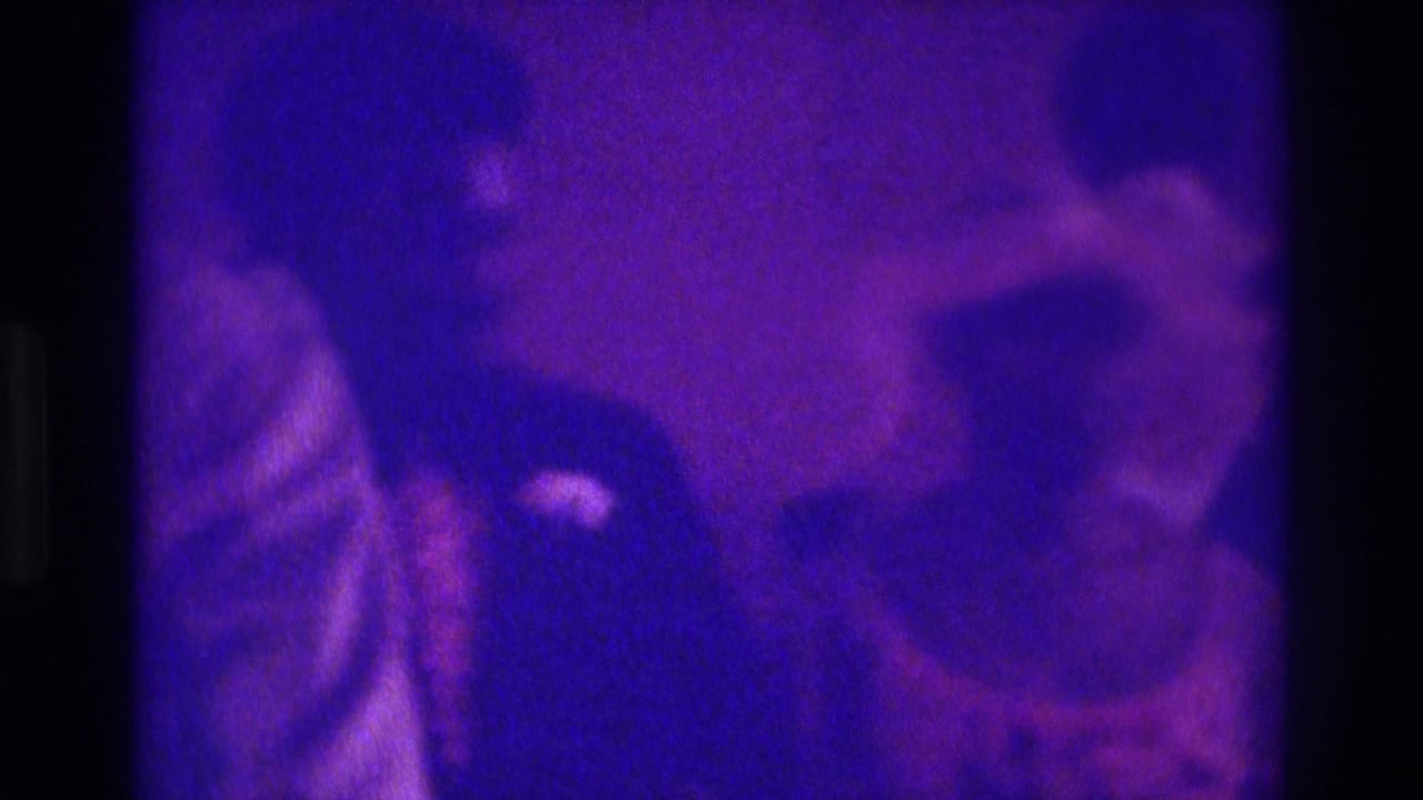 1975 Atlanta home-movie frame showing blurred guests in formal dress under deep purple indoor lighting.