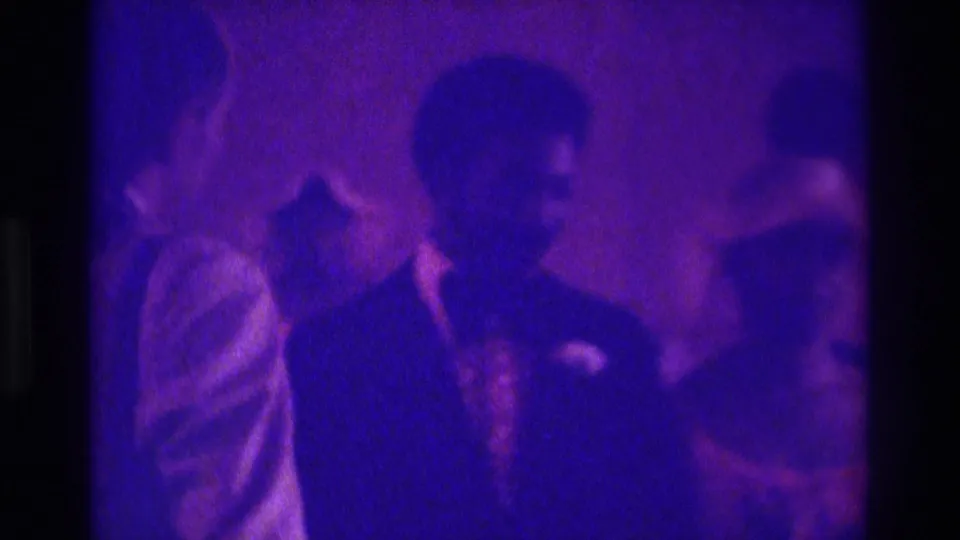 1975 Atlanta frame with a suited man and woman in a light dress standing in a crowded purple-lit interior.
