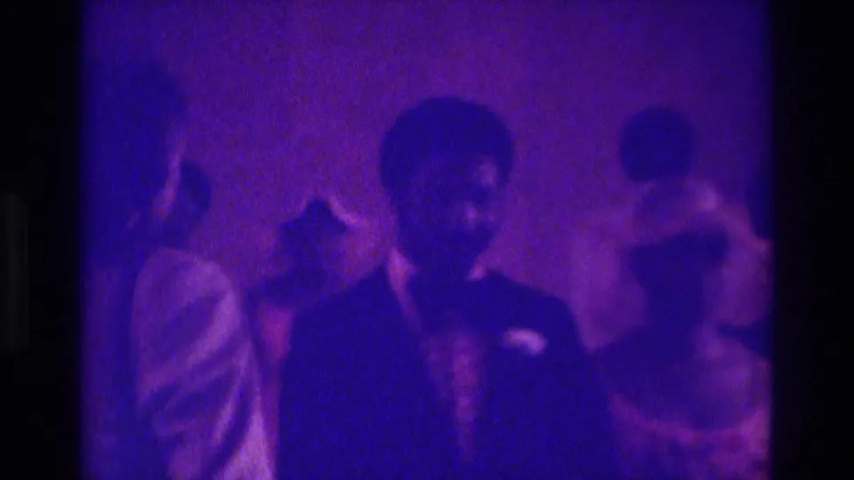 Soft 8mm frame from Atlanta in 1975 showing a man in a dark suit among formal guests in low purple light.