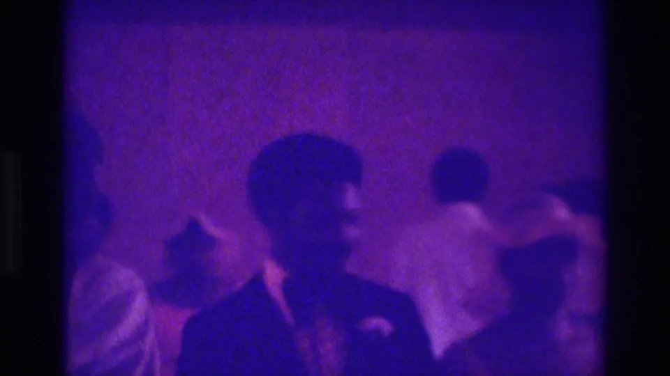 1975 Atlanta home-movie frame with a suited man in profile and guests shifting through a purple-lit indoor event.