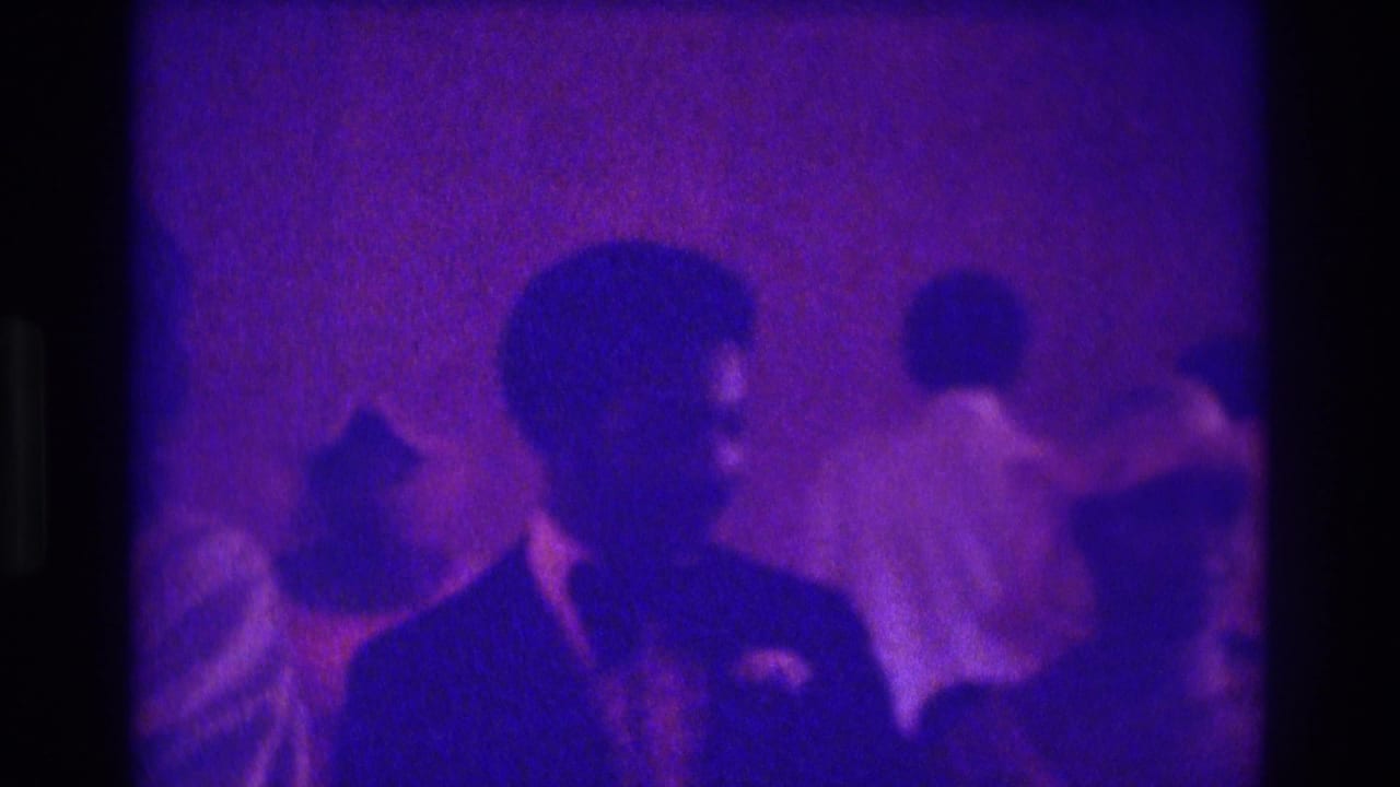 1975 Atlanta 8mm wedding footage shows a suited man with boutonniere near guests in a dim purple-tinted indoor ceremony space.