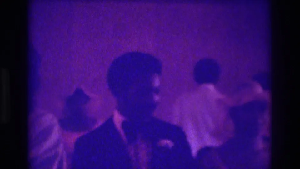 1975 Atlanta 8mm wedding footage shows a suited man with boutonniere near guests in a dim purple-tinted indoor ceremony space.