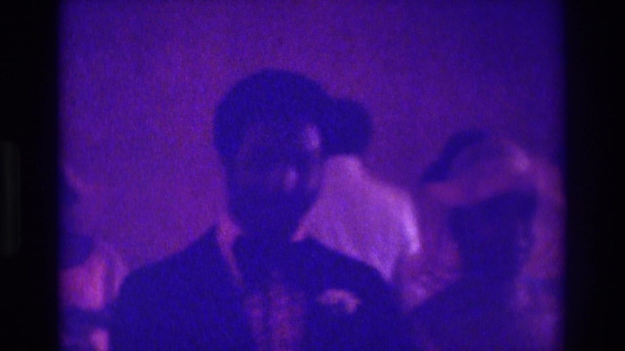 Suited man in foreground with guests and a woman at the side in purple light.