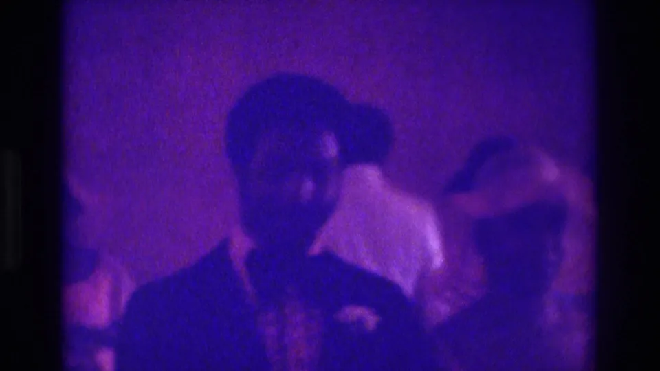 A suited man stands in the foreground of a 1975 Atlanta wedding scene while guests and a woman remain partly visible behind him.