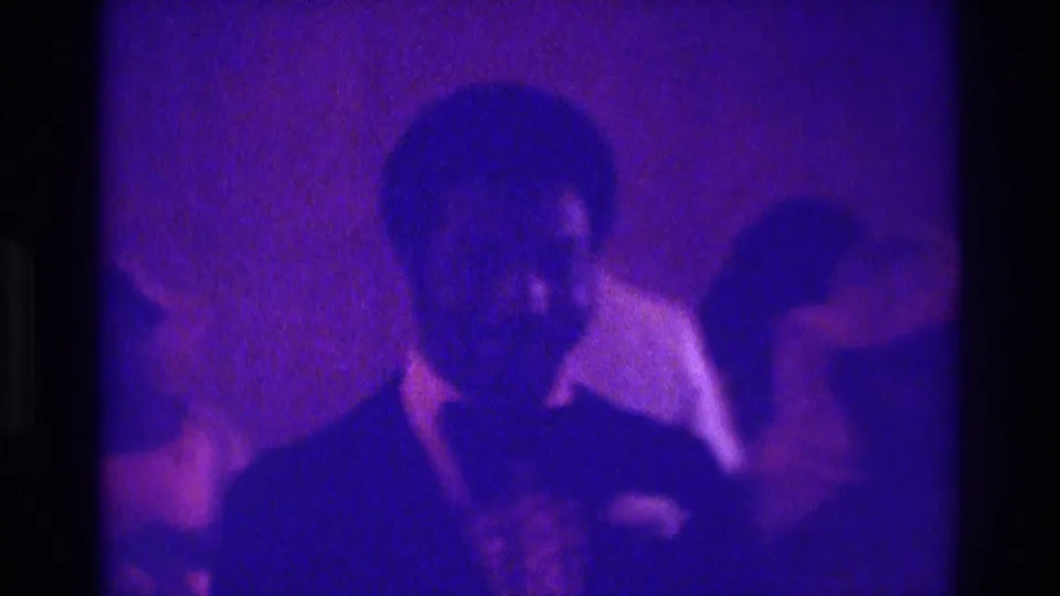 Soft 8mm footage from the 1975 Atlanta wedding shows a suited man close to camera with another guest in a light shirt behind him.