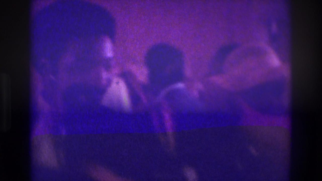 1975 Atlanta home-movie frame showing a dim wedding aisle with silhouetted figures and guests under a strong purple cast.