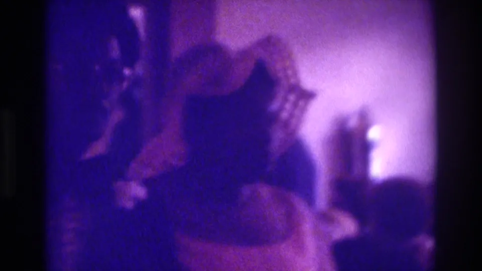 Purple-tinted 8mm frame from a 1975 Atlanta wedding, with a bride-like figure and headpiece visible among seated guests.