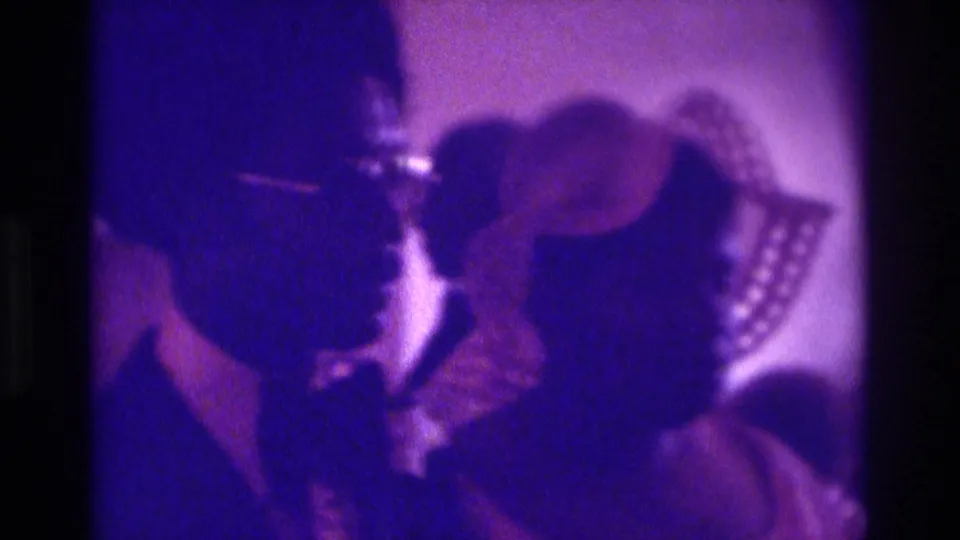 1975 Atlanta home-movie frame showing a tuxedoed figure near a bride-like figure, with guests seated in a dim wedding interior.