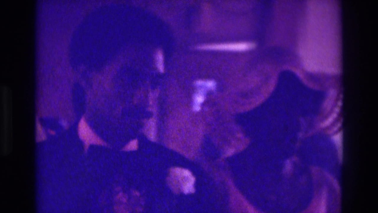 Dark-suited wedding party member with boutonniere beside guests in a purple-tinted interior.