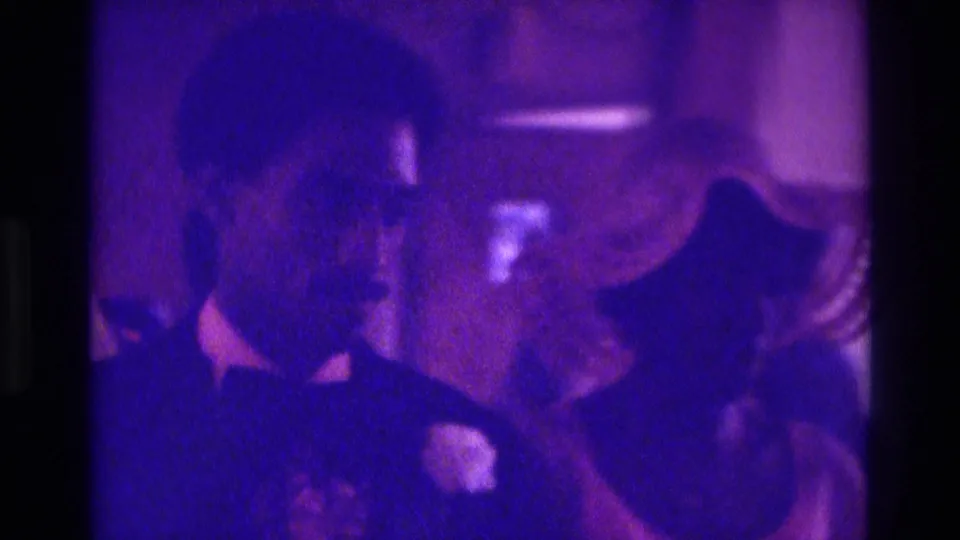 A wedding attendant in a dark suit and white boutonniere appears beside seated guests in 1975 Atlanta home-movie footage.