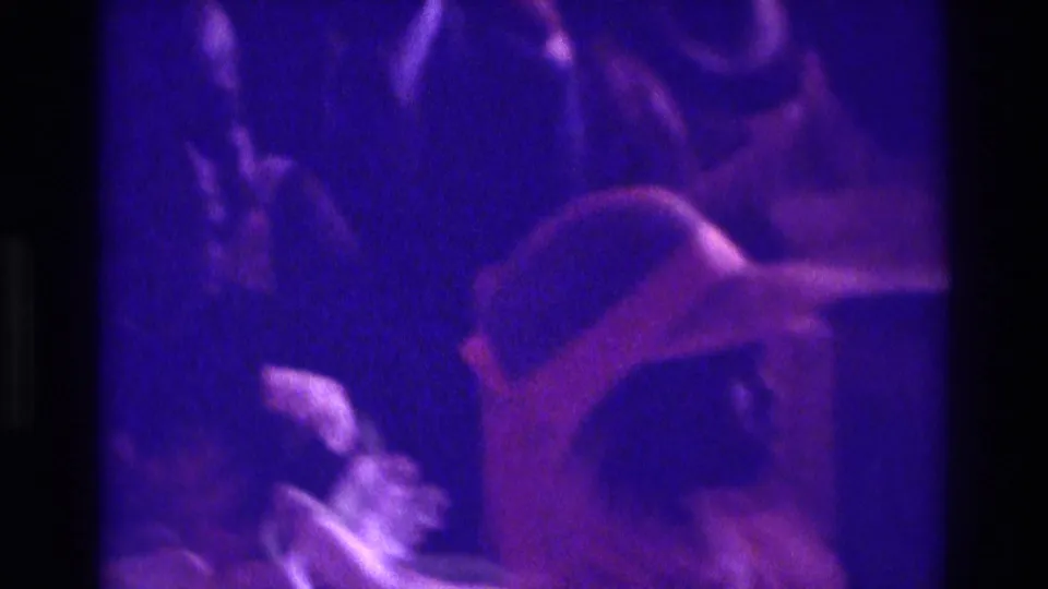 1975 Atlanta 8mm frame showing a dim indoor formal gathering with a brimmed hat, partial faces, and heavy purple film color shift.