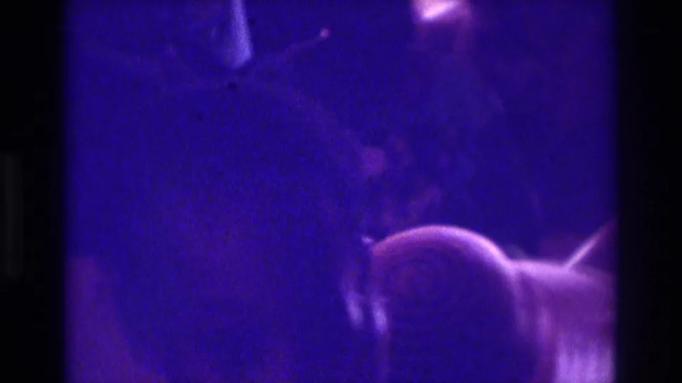 1975 Atlanta 8mm frame showing blurred indoor figures, a brimmed hat, and dark film-frame edges in strong purple color.