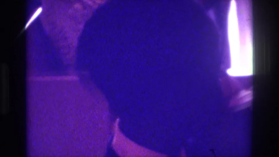1975 Atlanta home-movie frame with a dark head-and-shoulder silhouette, bright vertical light, and purple 8mm grain in a dim interior.