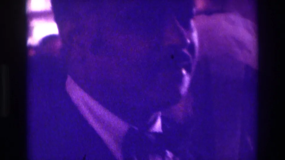 1975 Atlanta home-movie frame showing a veiled bride and escort partly obscured by purple-blue ceremony lighting.