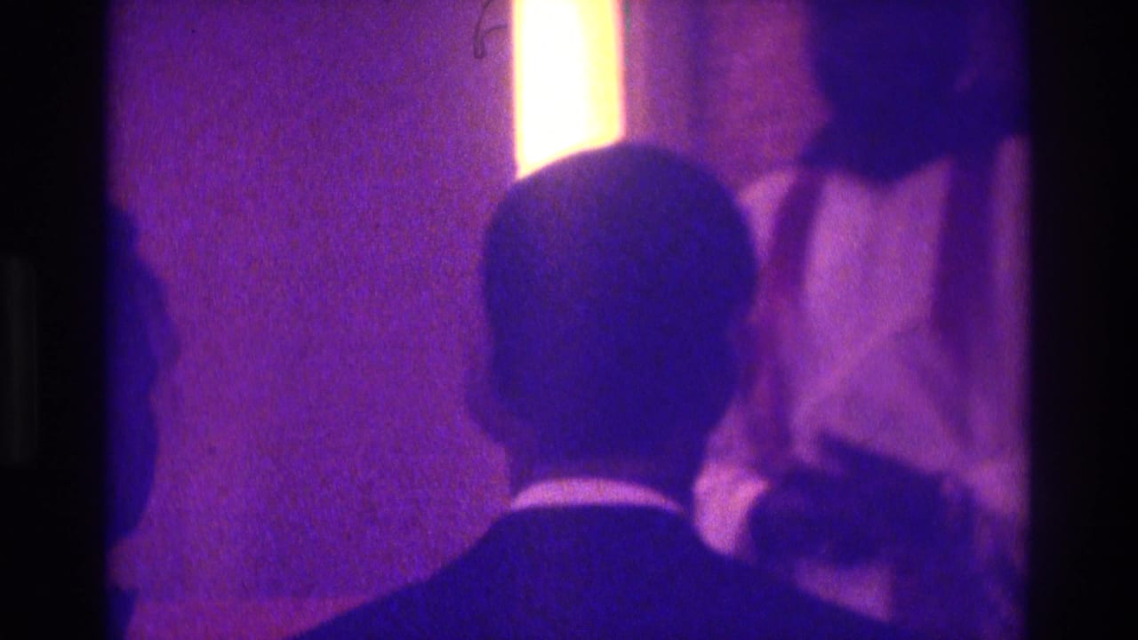 Atlanta 1975 home-movie frame showing the backs of formally dressed attendees watching a dimly lit religious ceremony.