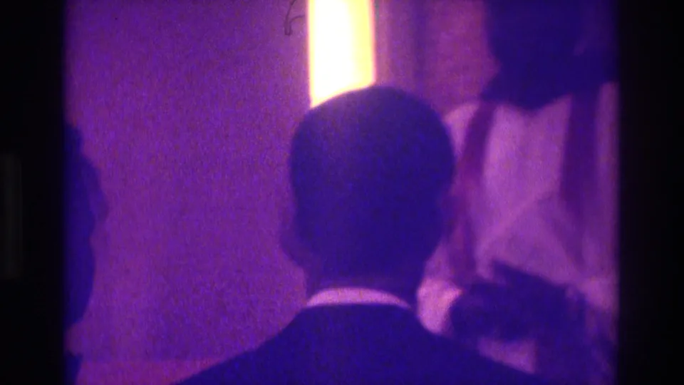 Atlanta 1975 home-movie frame showing the backs of formally dressed attendees watching a dimly lit religious ceremony.