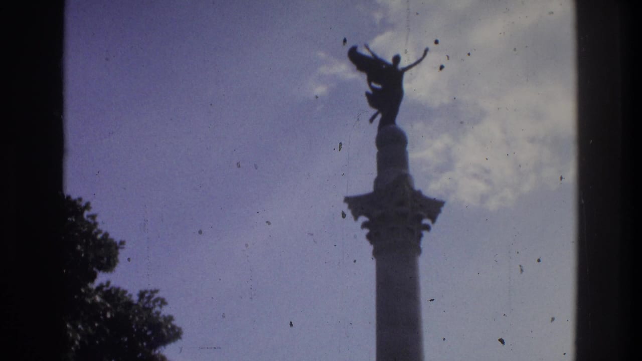 1975 Atlanta 8mm frame showing a tall monument column and winged statue silhouetted above trees and a cloudy blue sky.