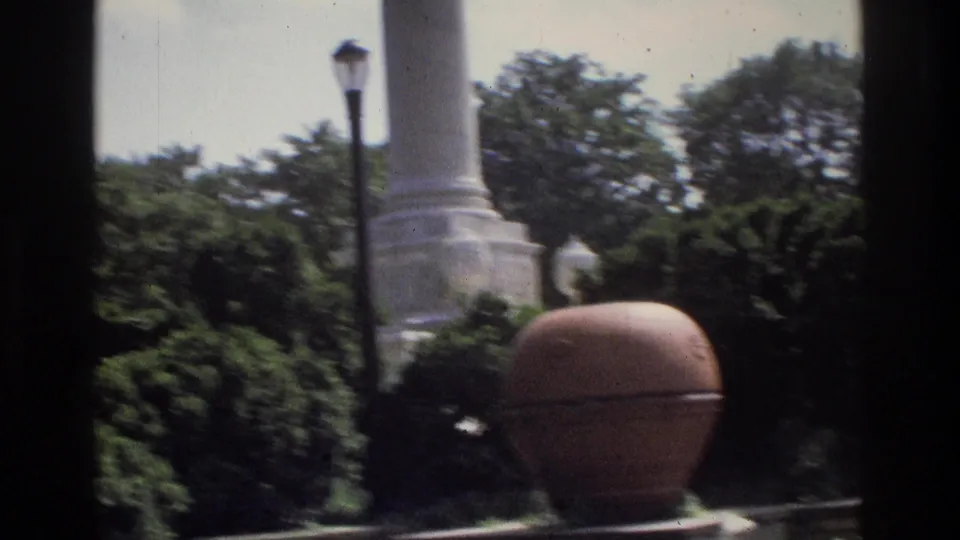 1975 Atlanta 8mm frame showing the lower monument, garden shrubs, lamp post, trees, and a round planter in sunlight.