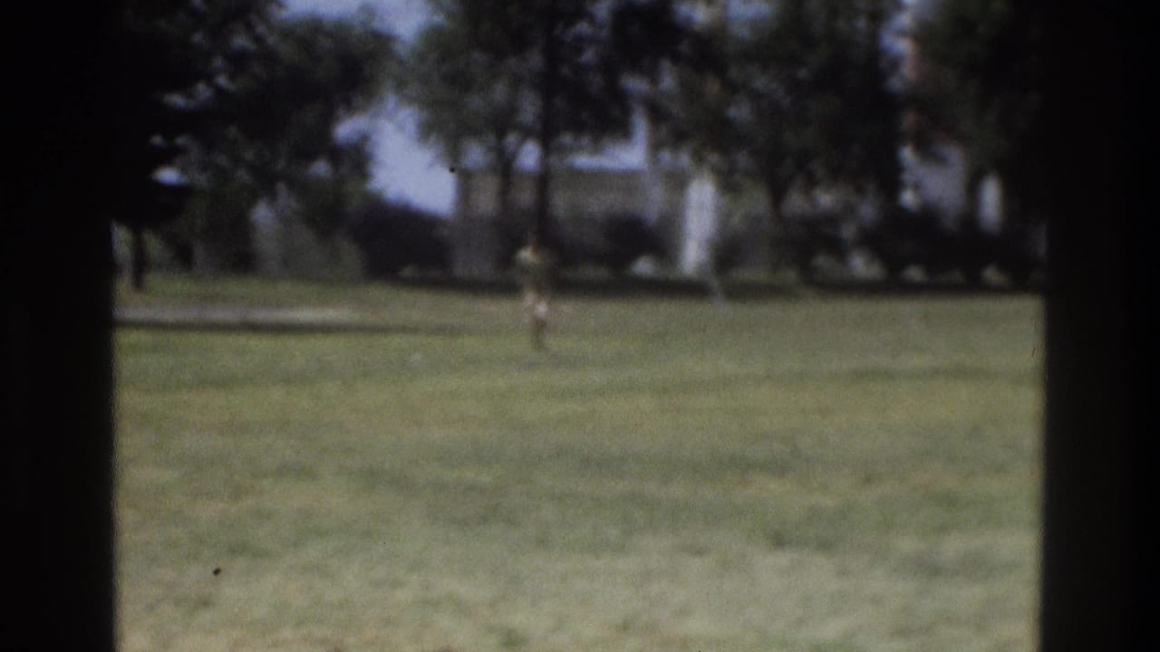 1975 Atlanta home-movie frame showing a distant runner on a lawn, seen through a dark window-like edge.