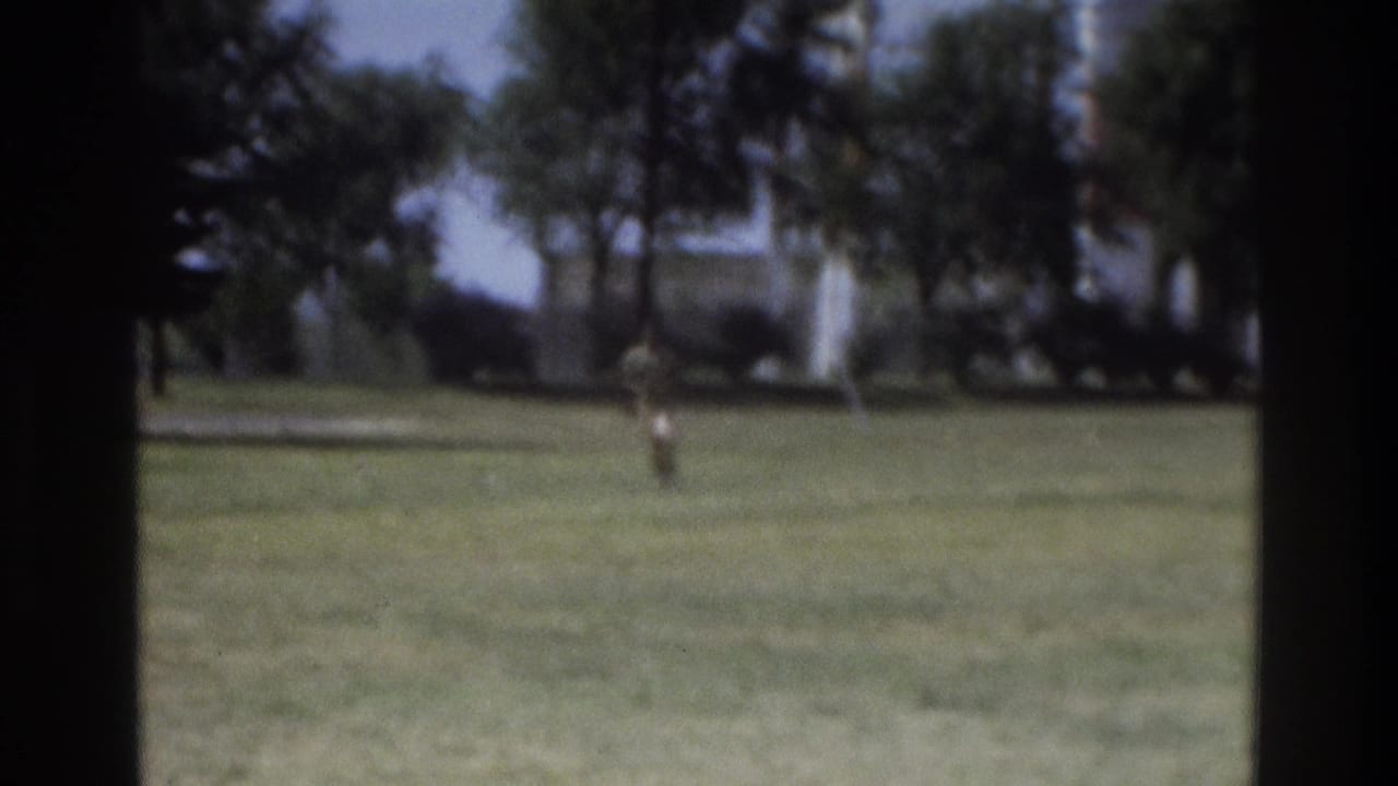 Soft 8mm frame of a person running across a lawn in Atlanta, with trees and a distant structure behind them.
