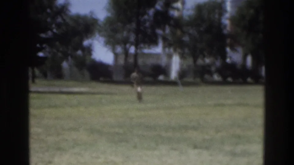 Soft 8mm frame of a person running across a lawn in Atlanta, with trees and a distant structure behind them.