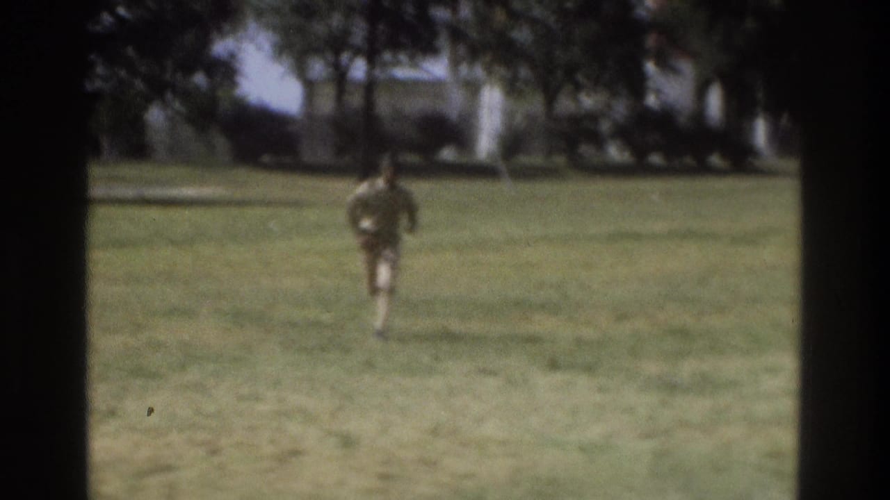 Black uniformed man running across a grassy Atlanta field in a soft 1975 8mm home-movie frame.