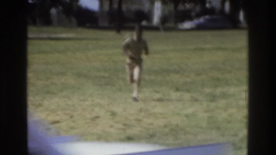 1975 Atlanta home-movie frame of a uniformed runner approaching as a car briefly crosses the foreground.